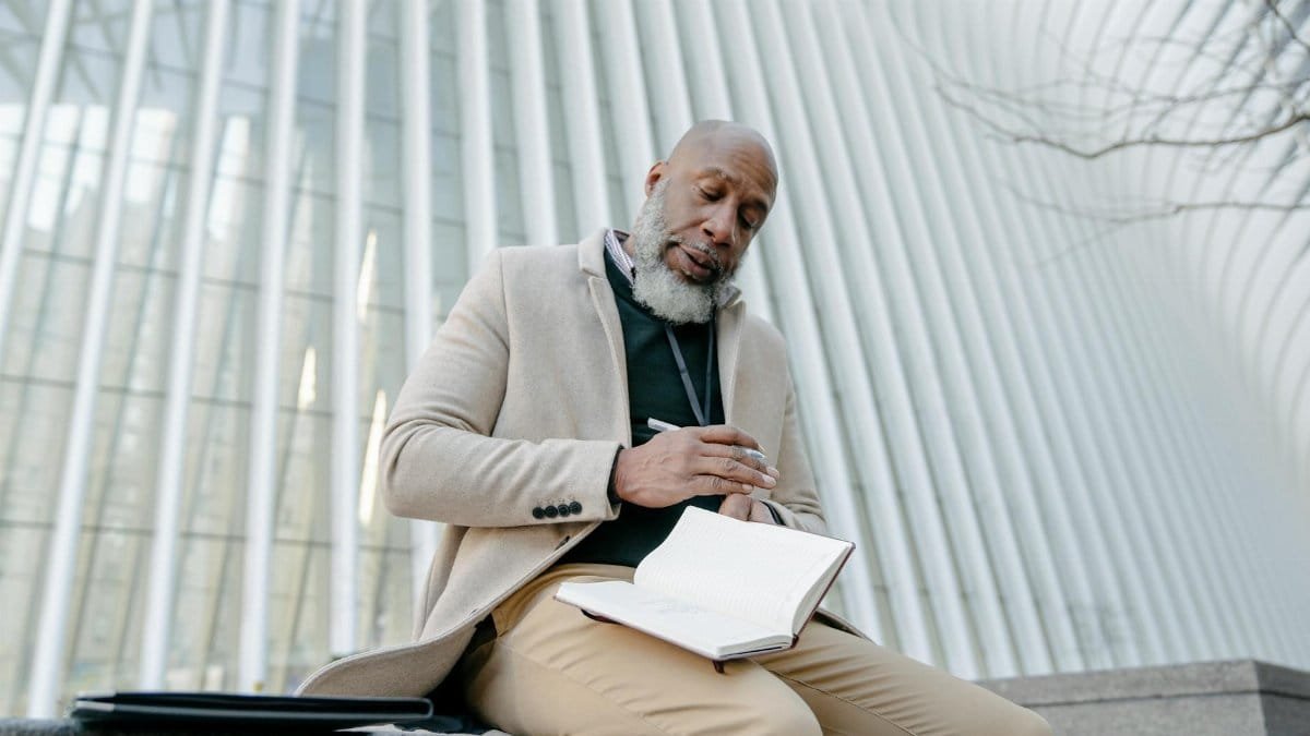 Mature bald man in beige coat writing notes on a bench outside modern architecture.