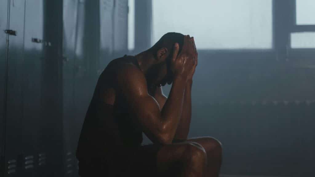 Sweaty man resting on bench in dimly lit locker room, showing exhaustion and recovery.
