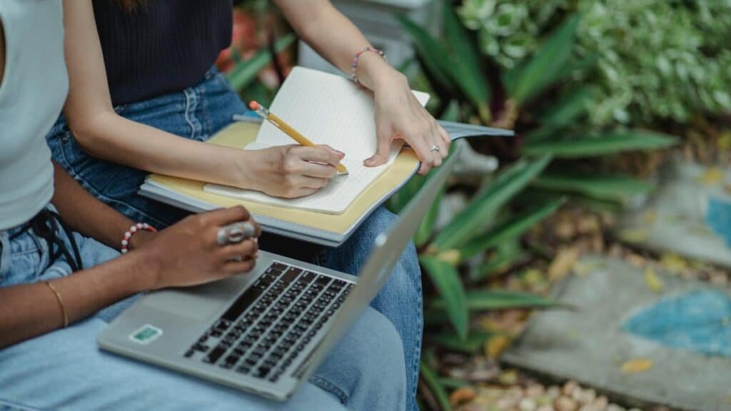 Crop faceless multiracial students in blue jeans using laptop and taking notes in workbook while preparing for exam together in lush summer park