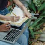 Crop faceless multiracial students in blue jeans using laptop and taking notes in workbook while preparing for exam together in lush summer park