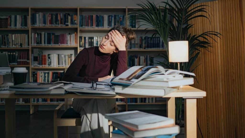 A tired woman surrounded by books, studying in a library, feeling overworked and stressed.