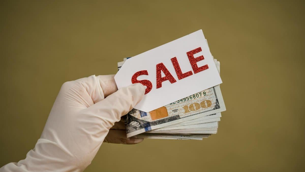 A hand wearing latex gloves holds a stack of dollar bills with a sale sign, symbolizing discount or cash transaction.