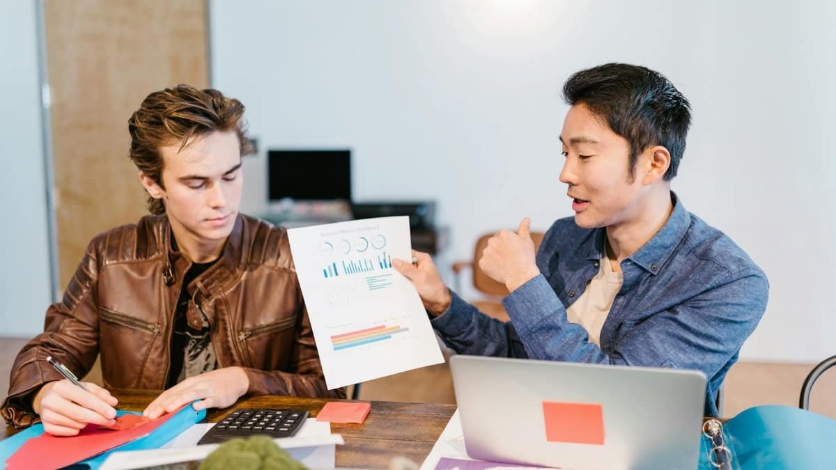 Two men discuss graphs and data at a shared workspace in a modern office setting.