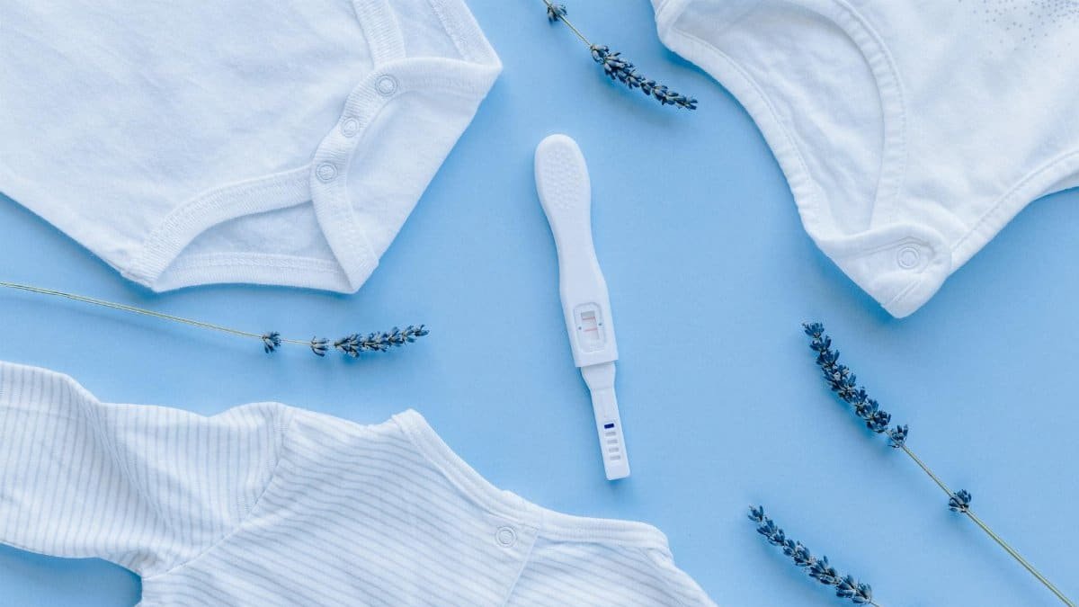 Flat lay of baby clothes and positive pregnancy test on blue background.