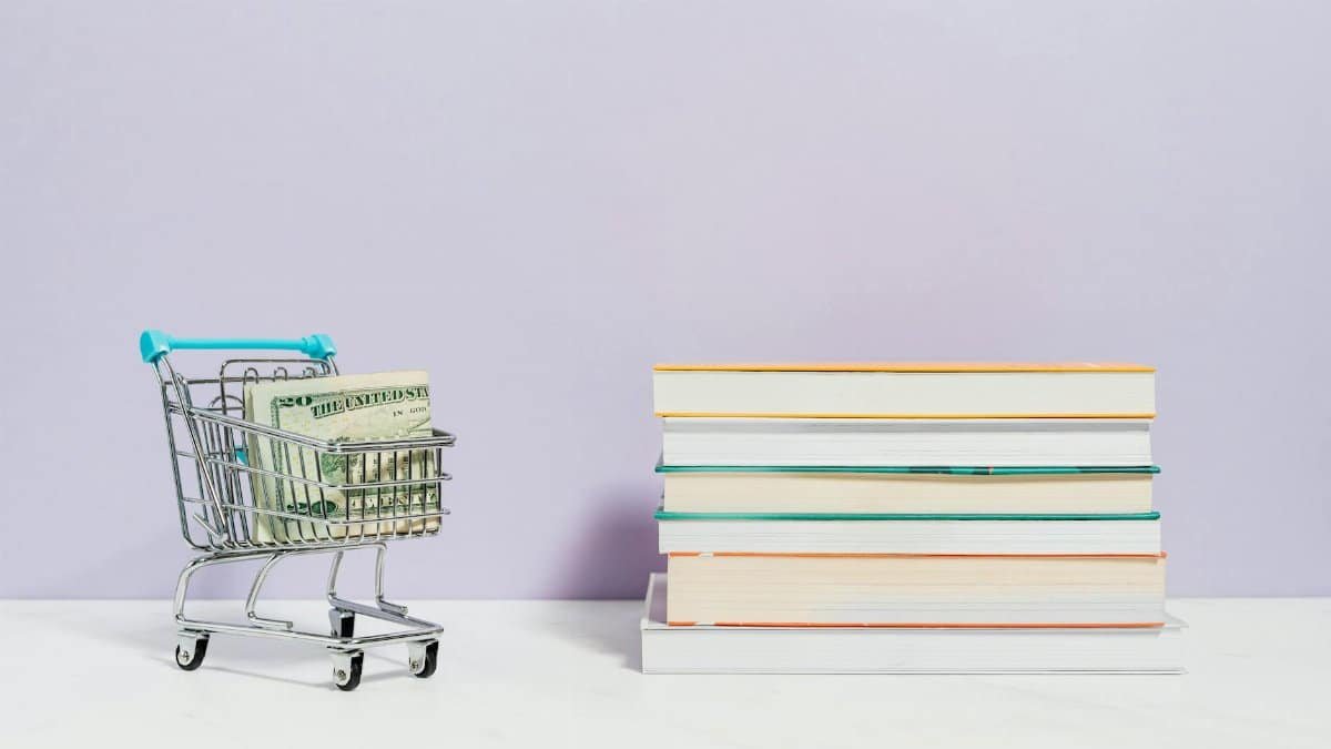 A shopping cart with dollar bills next to a stack of textbooks on a light background.
