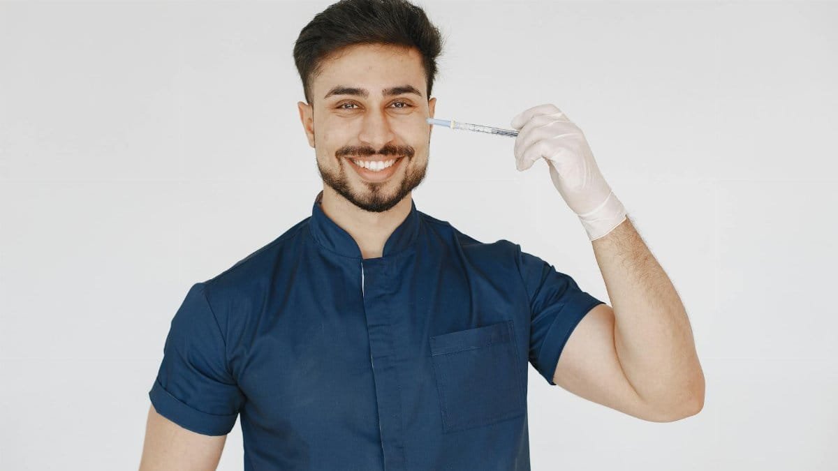 A confident doctor in blue scrubs smiling and holding a syringe in a studio setting.