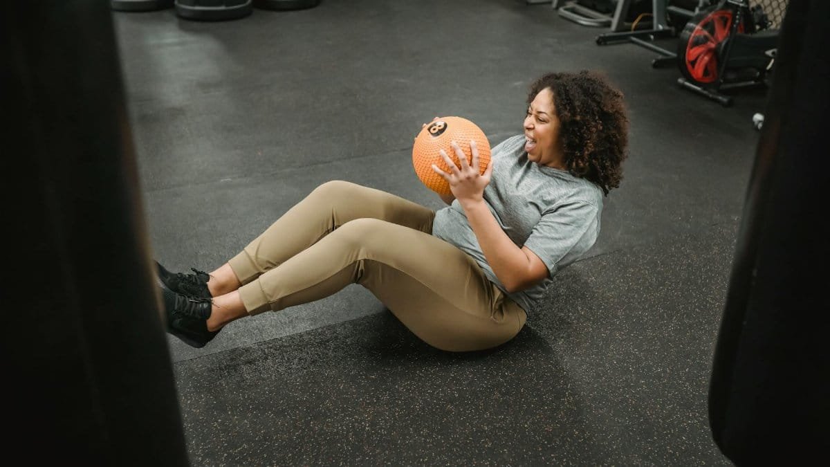 Plus-size woman doing an intense workout with a medicine ball in a gym setting.