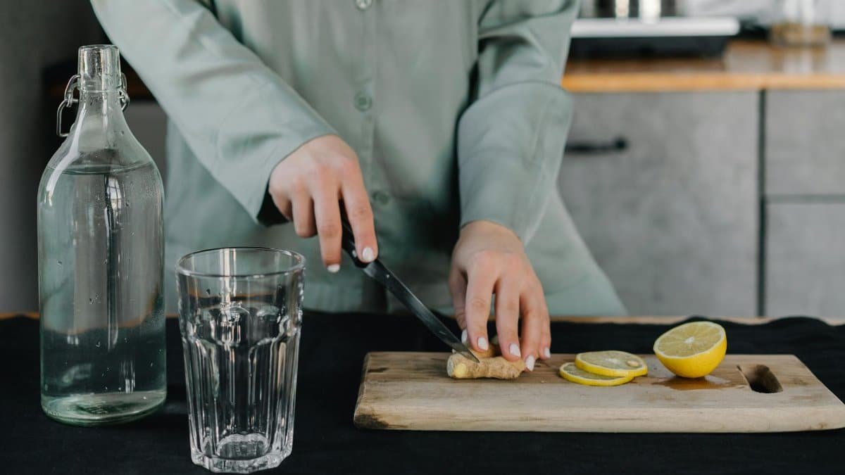 Hands cutting ginger next to lemons, a glass, and water bottle on a wooden board indoors.