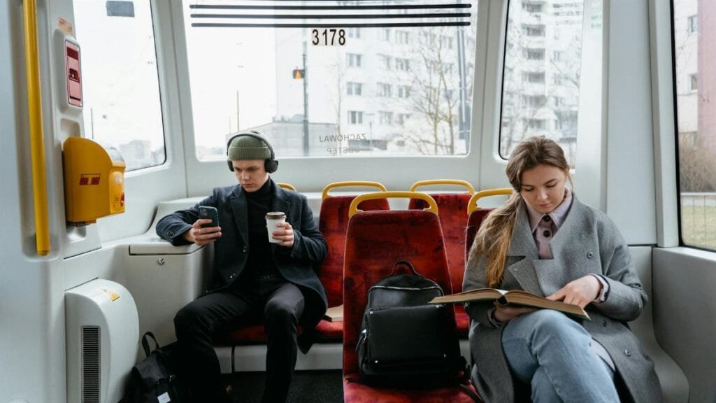 Two commuters on a tram, using gadgets and reading, showcasing a typical urban lifestyle.