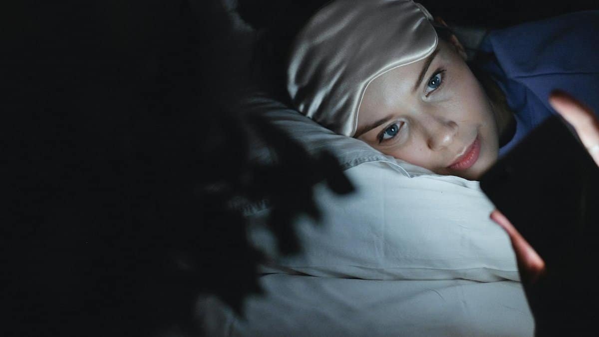 A woman in bed wearing a sleep mask uses her phone at night, creating a cozy ambiance.