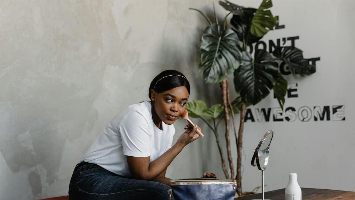 A young woman applies makeup in a cozy, stylish home setting with a mirror and indoor plants.