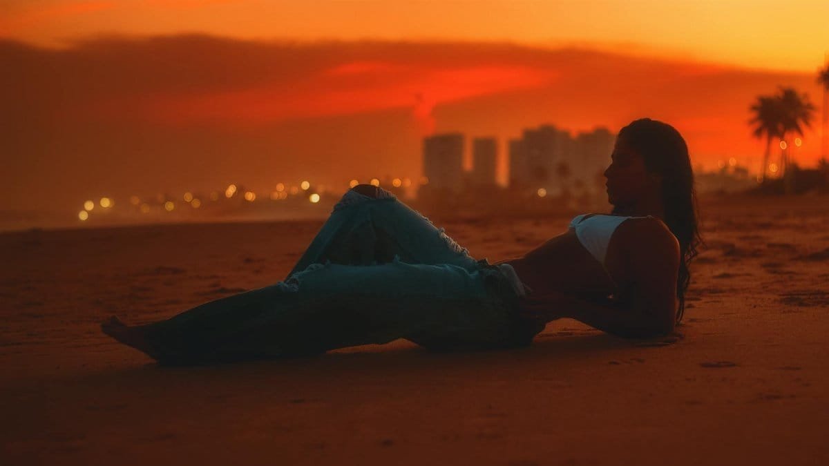 Silhouette of a woman lying on the beach during a vibrant sunset, with a cityscape in the background.