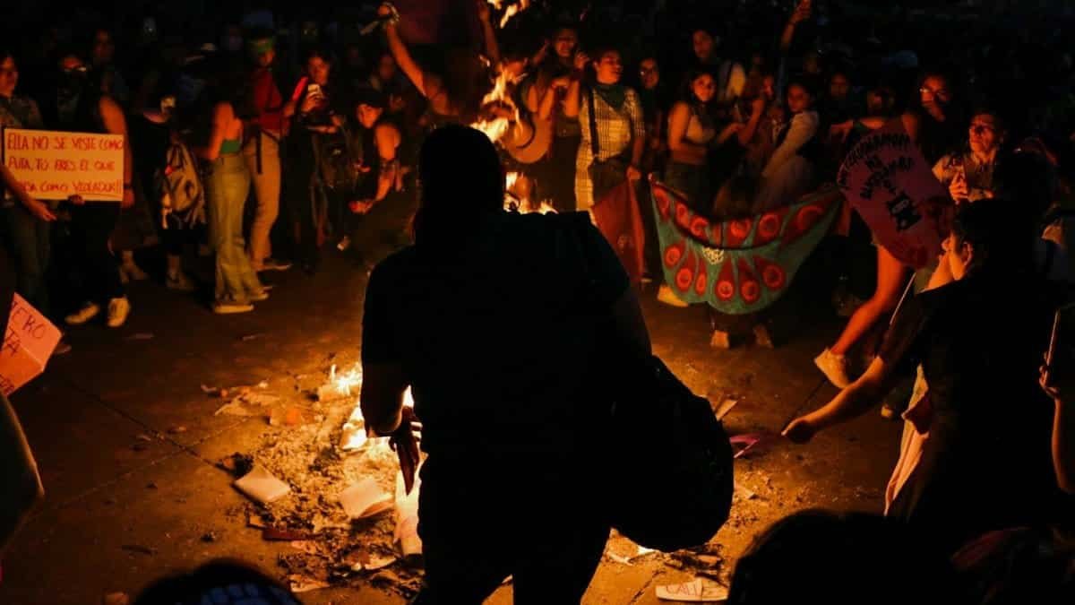 A large group of protestors in Mexico City gathered around a burning pile, holding signs during night protest.