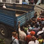 A diverse group of people boarding a blue truck in a rural area, illustrating travel and community.