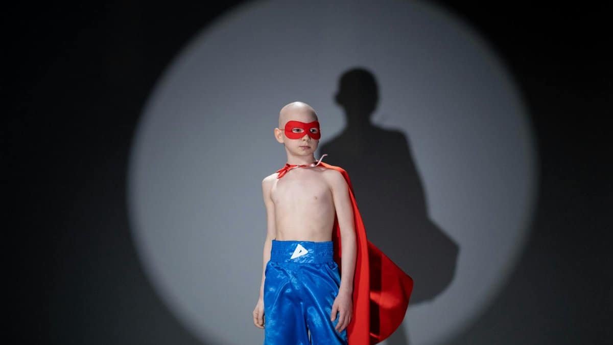 A child in a superhero costume poses confidently in a studio spotlight.