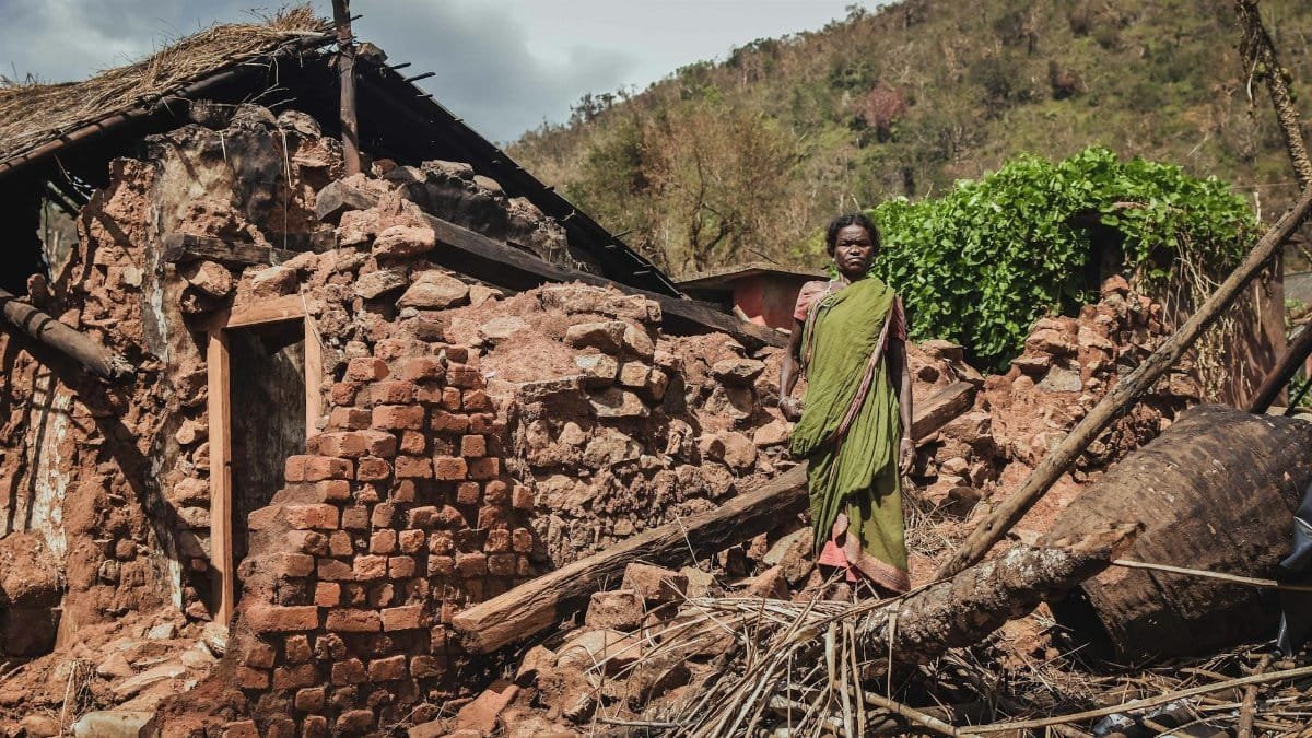Woman stands in front of a destroyed house after a disaster in rural India, depicting struggle.