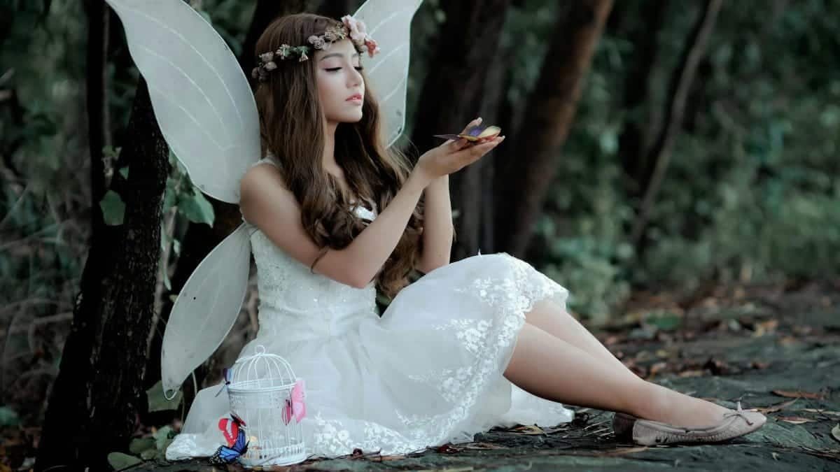 A captivating image of a fairy in white dress with wings, sitting in a woodland setting, holding a butterfly.