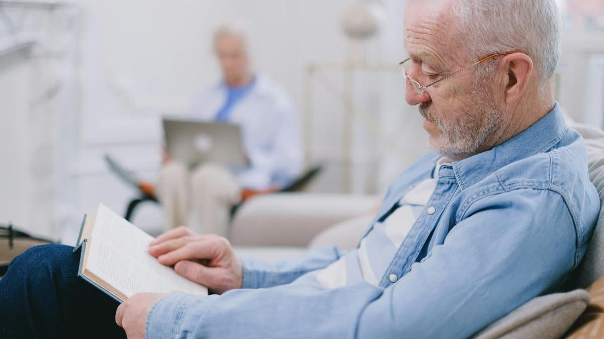 Elderly man with eyeglasses reading a book indoors while another senior uses a laptop.