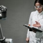 Researchers in lab coats and safety glasses engaging with a robotic arm in a lab setting.