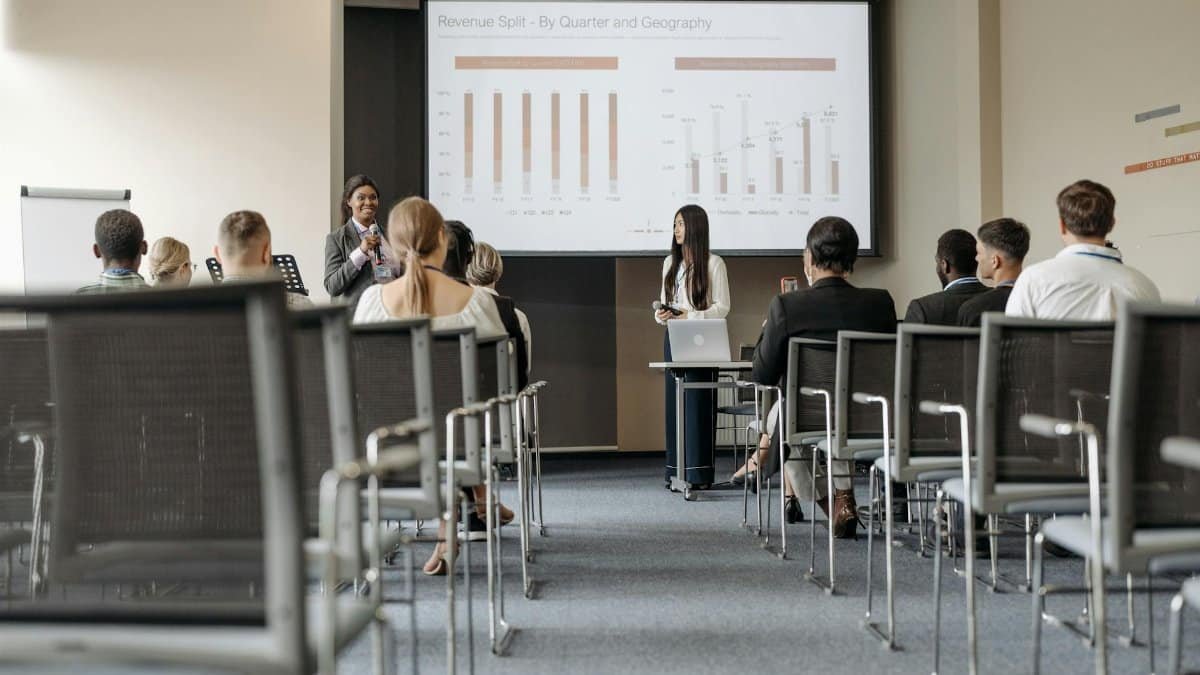 Business conference attendees listen to a presentation on revenue split by quarter and geography.