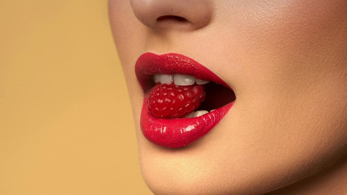 Vibrant red lips holding a fresh raspberry in sensual close-up shot.