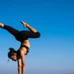 A woman performing a yoga handstand on a rock by the sea under a clear blue sky.