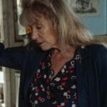 A thoughtful senior woman leans on a refrigerator, evoking emotion and nostalgia in a cozy kitchen setting.