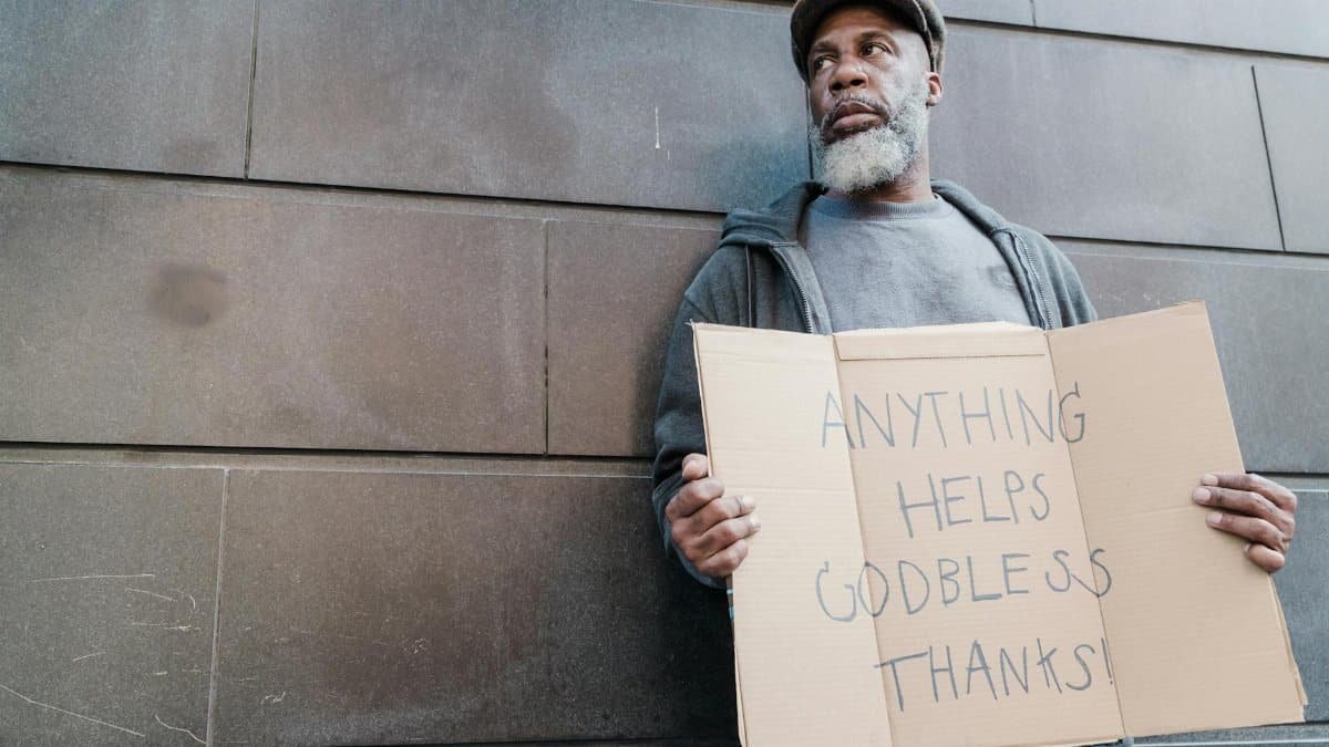 Elderly man with beard holding a handwritten sign seeking help and blessings against a stone wall.