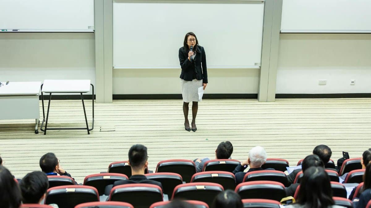 Female speaker presenting to an audience in a modern auditorium setting.