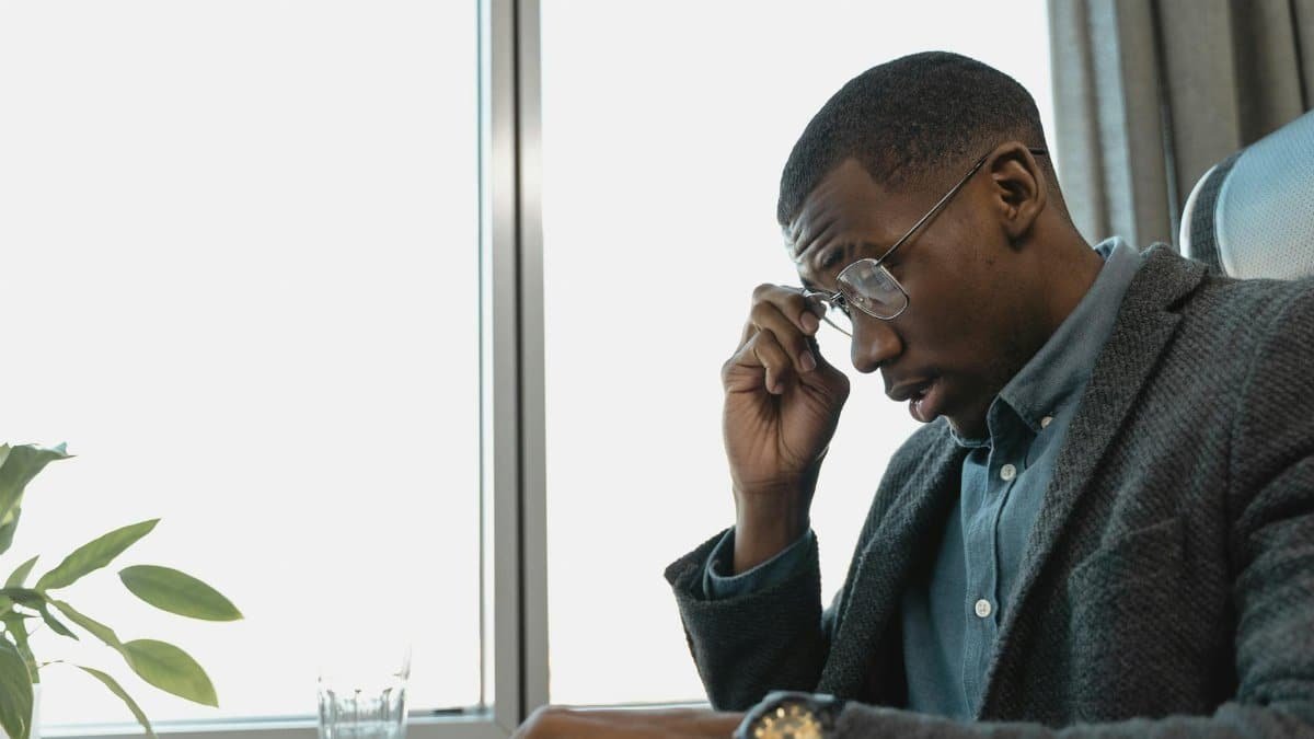 Exhausted businessman in glasses contemplating at office desk near window.