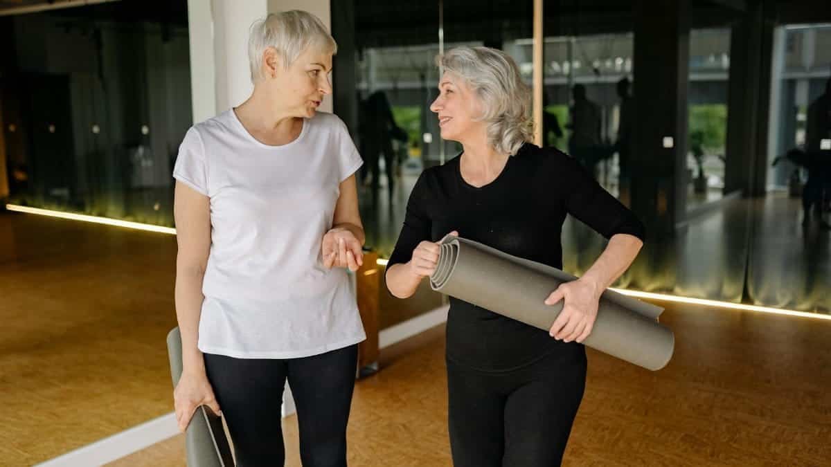 Two senior women engaging in conversation while holding yoga mats inside a fitness studio.