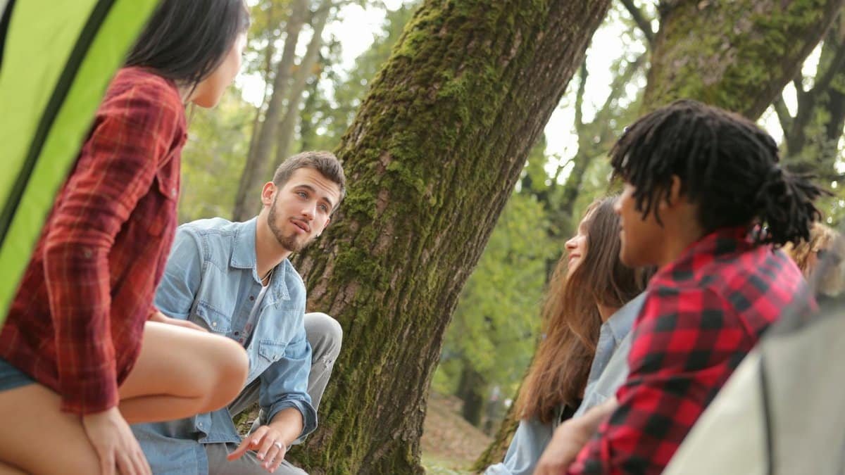 Group of young people in casual clothes sitting on plaid around near trees with moss while talking and looking at each other