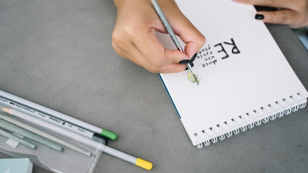 Close-up of hand lettering 'Recycle' on a notepad using drawing materials, emphasizing eco-friendly practices.