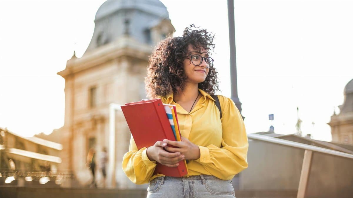 Smiling female student with curly hair and glasses holding books on campus steps.