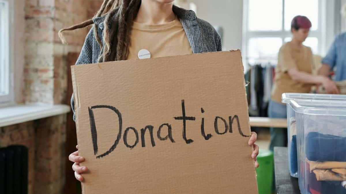 Volunteer holding a cardboard donation sign in a community center with clothing and supplies.