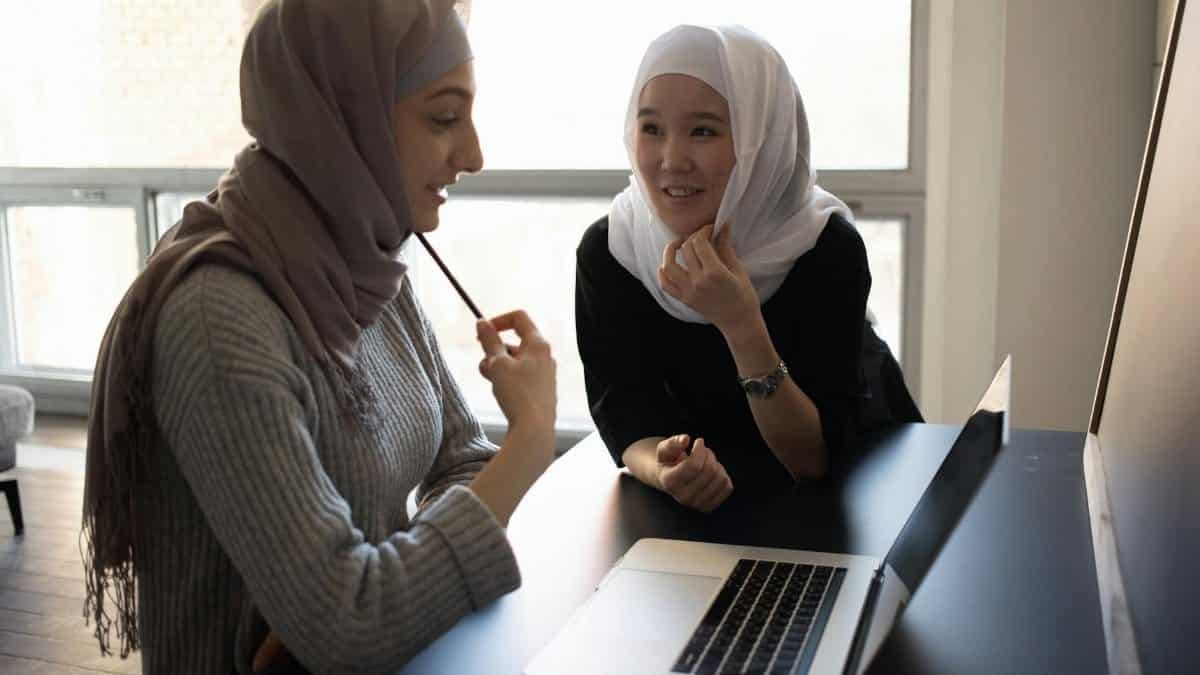 Positive multiethnic Muslim female students in traditional headscarves sitting at table and surfing netbook while preparing for lesson together in university