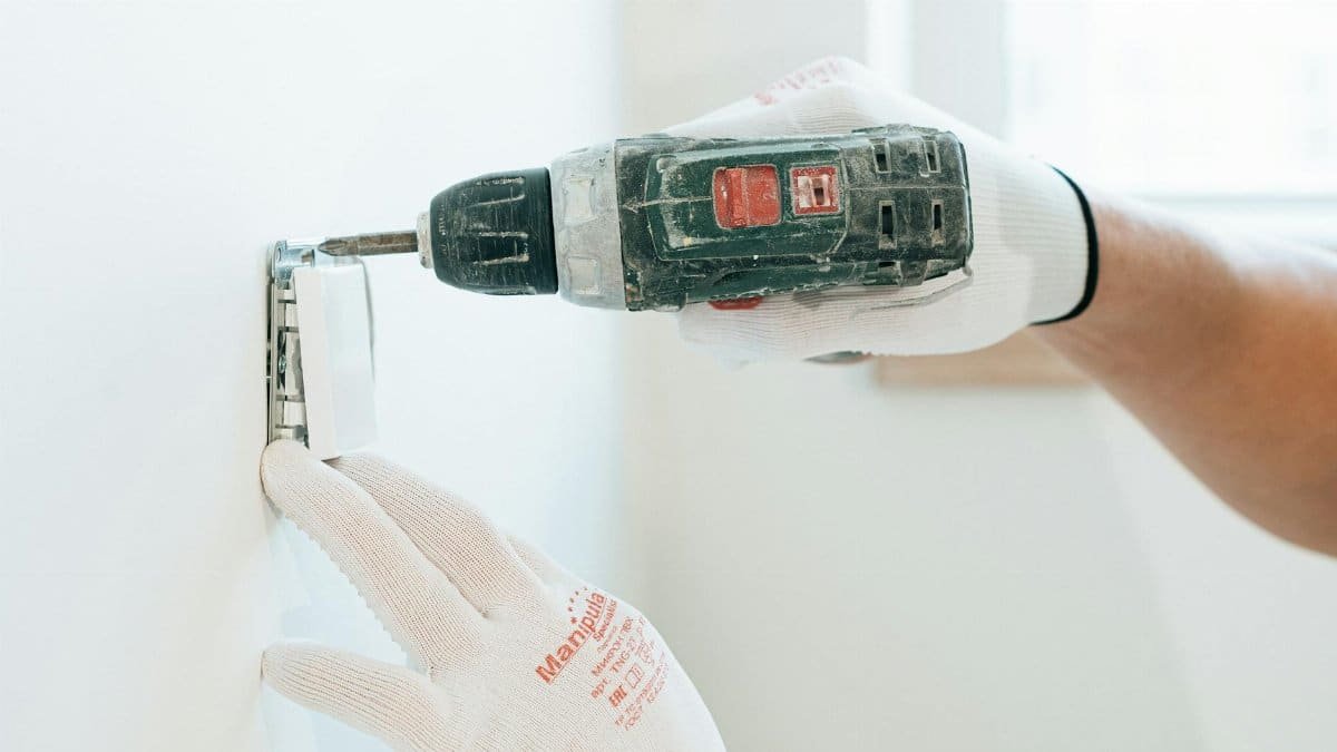 Gloved hands using a drill to install a power outlet on a wall, focusing on precision and safety.
