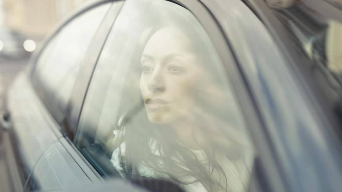 A woman reflects while sitting inside a car, looking through the window with a thoughtful expression.
