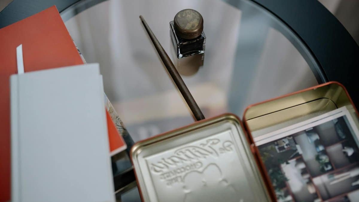 A close-up view of a vintage desk setup featuring notebooks, an ink bottle, and photos on a glass table.
