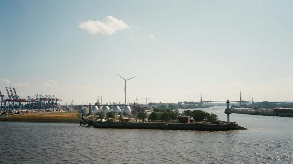 A scenic view of an industrial area by a river, featuring a wind turbine and bridges under a blue sky.