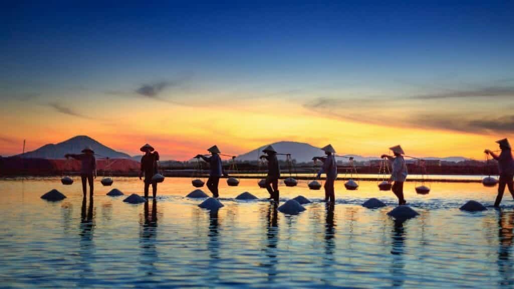 Silhouettes of salt farmers at sunset, reflecting on water with mountains in the background.
