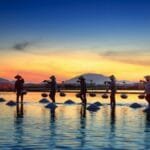 Silhouettes of salt farmers at sunset, reflecting on water with mountains in the background.