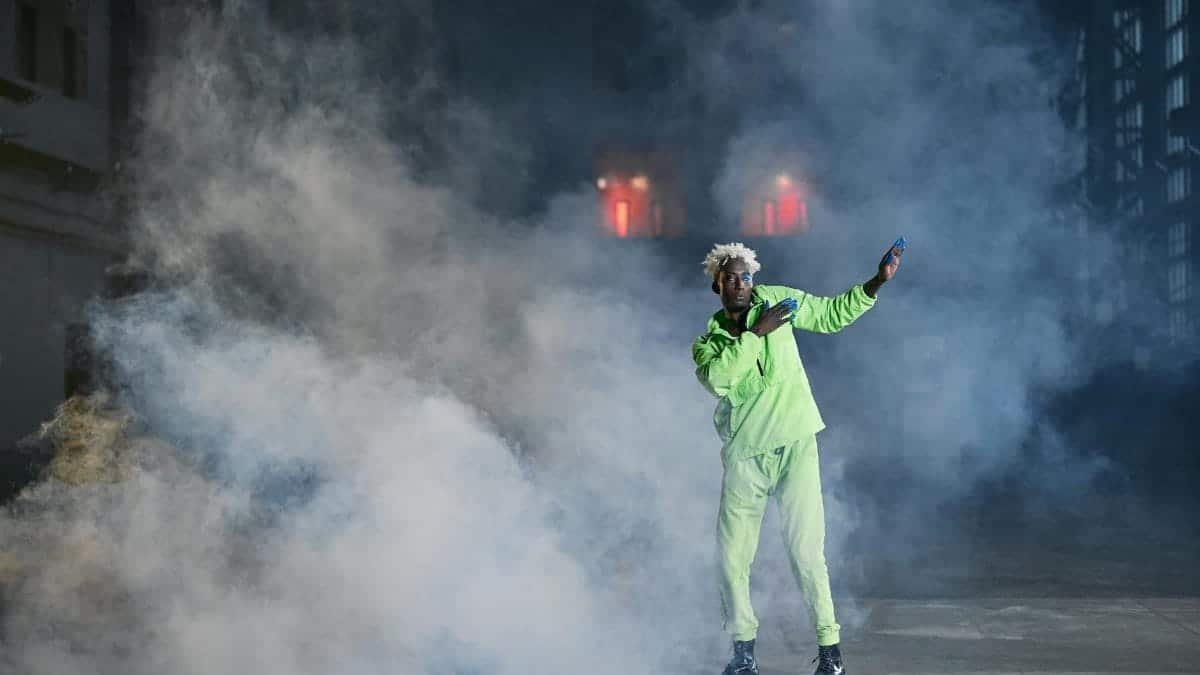 A performer stands on stage in a bold green suit surrounded by dramatic smoke effects, exuding energy.