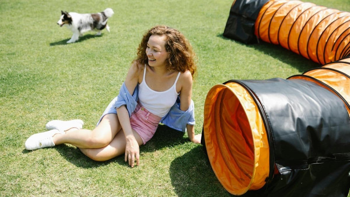 From above of laughing female with long curly hair sitting on green grass near playing tunnel while walking dog on sunny day in summer