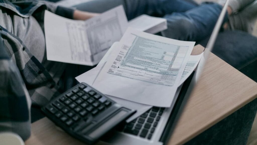 A young adult using a calculator and laptop to manage finances at home, surrounded by papers.