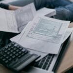 A young adult using a calculator and laptop to manage finances at home, surrounded by papers.