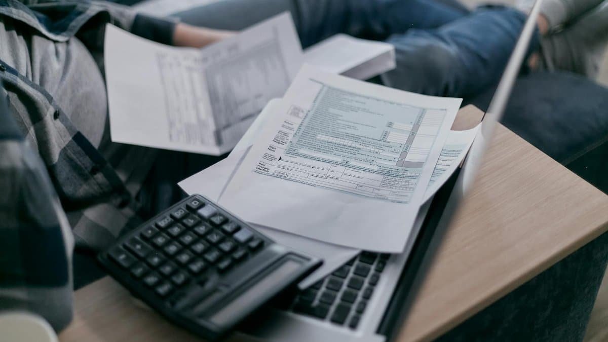 A young adult using a calculator and laptop to manage finances at home, surrounded by papers.