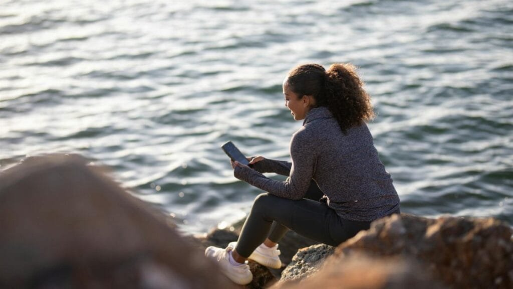 A woman sitting on a rocky shore using a smartphone, enjoying the serene seaside view.