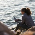 A woman sitting on a rocky shore using a smartphone, enjoying the serene seaside view.
