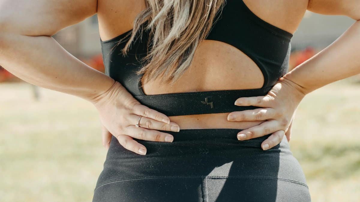 A woman in activewear holds her lower back, indicating muscle strain, outdoors.